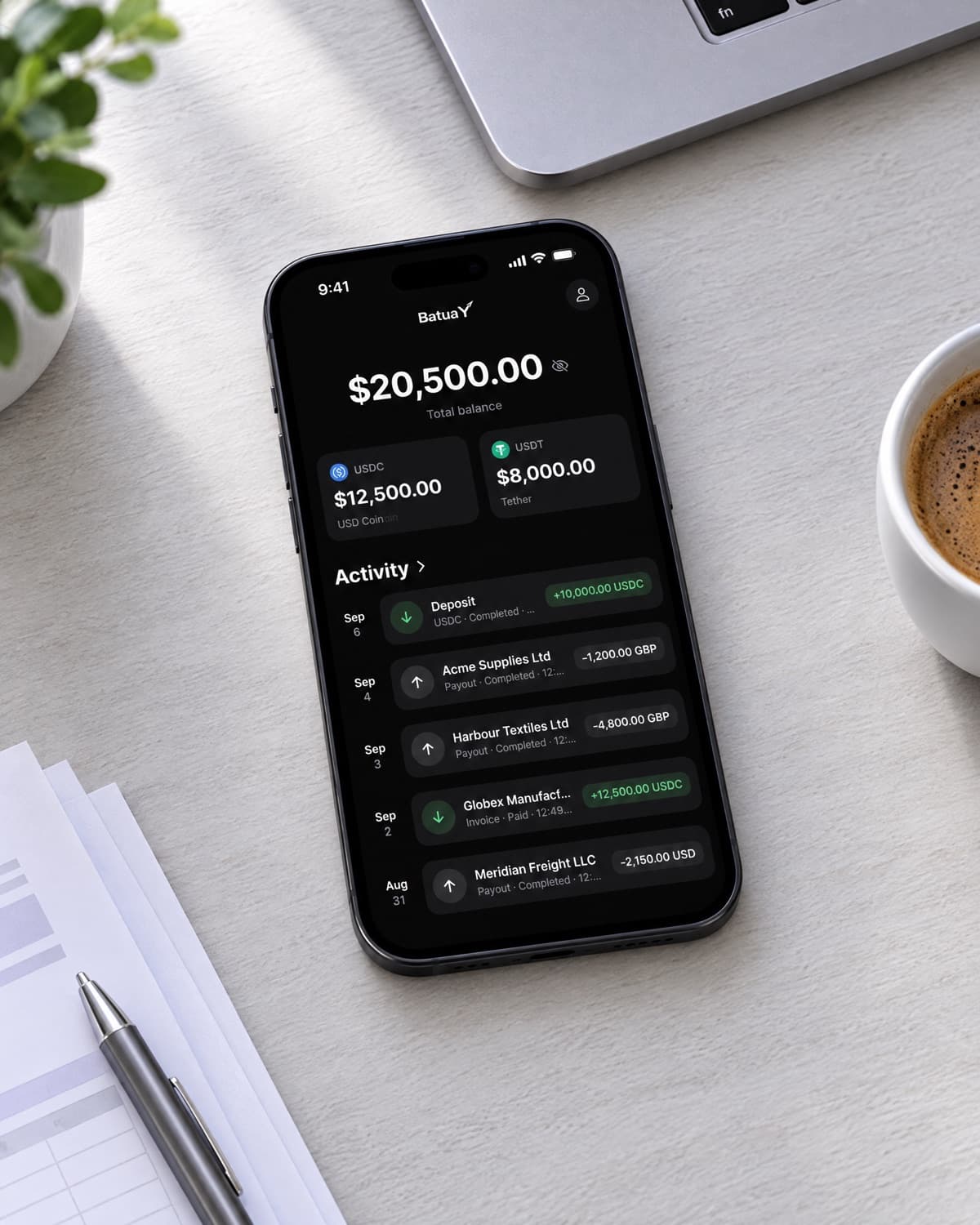 A phone on an office desk showing the BatuaY business balance with USDC and USDT and recent supplier payouts