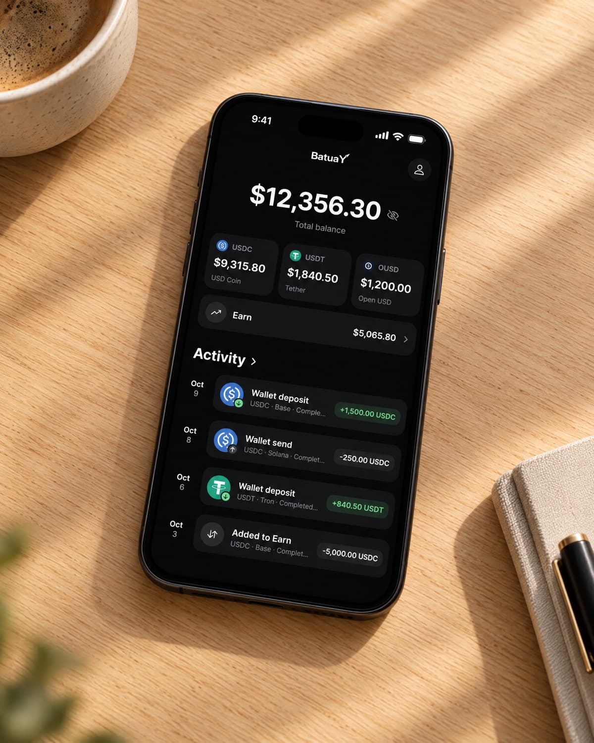 A phone on a wooden table showing the BatuaY home screen with USDC, USDT and OUSD balances